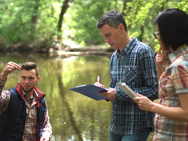 Three people taking water samples and writing notes on edge of body of water