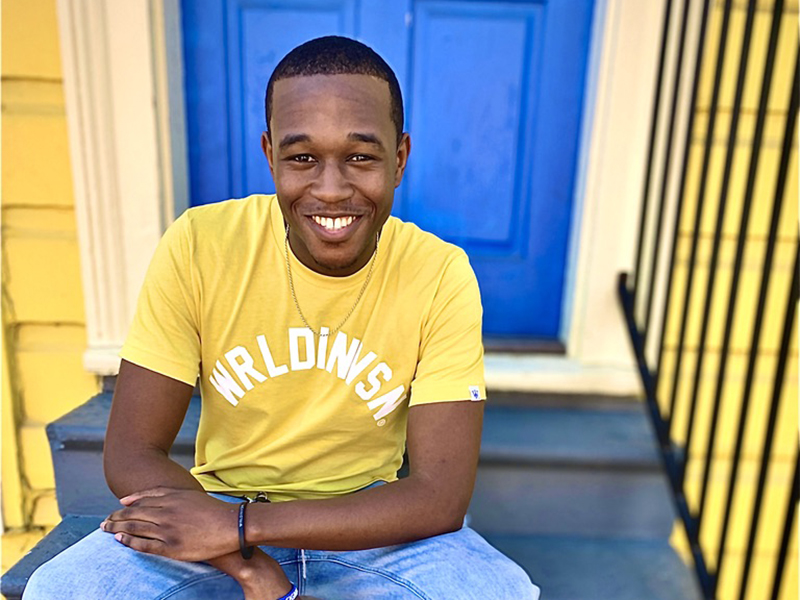 Photo of Daniel Triggs in yellow shirt sitting on the steps of a yellow home
