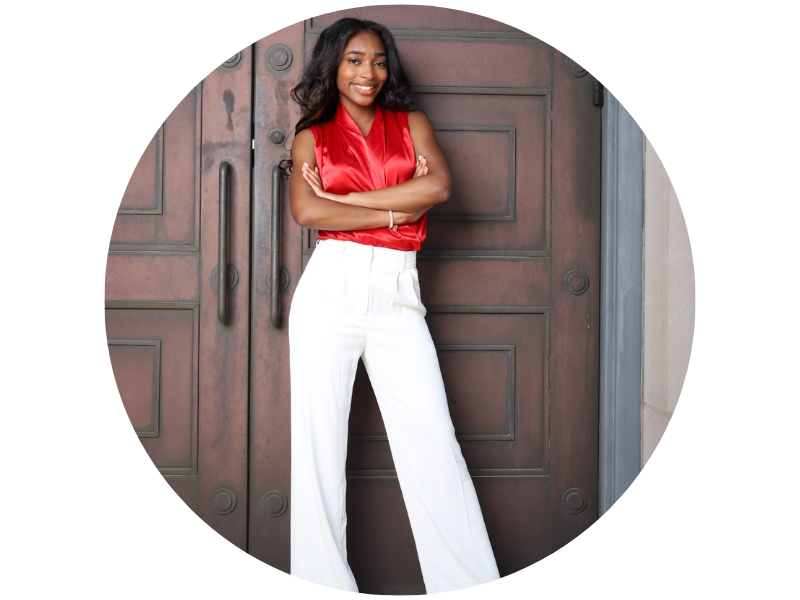 Bryson Reems, standing, arms crossed in front of a set of wooden doors, wearing a red blouse and white pants, looking directly at camera