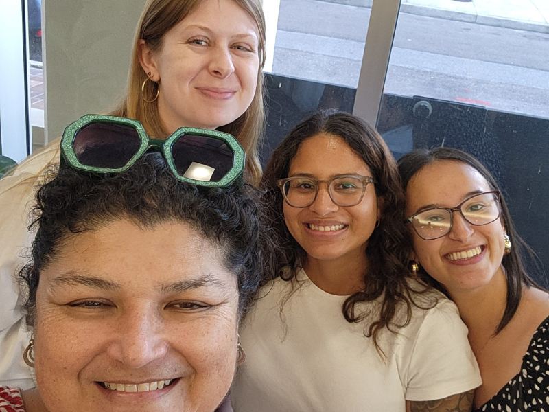 Four women smiling together indoors near a window.