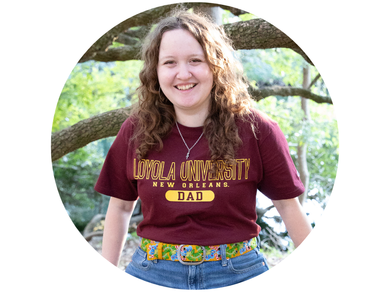Maeve Karnes, smiling, wearing a Loyloa University t-shirt, sitting on large tree branch. 