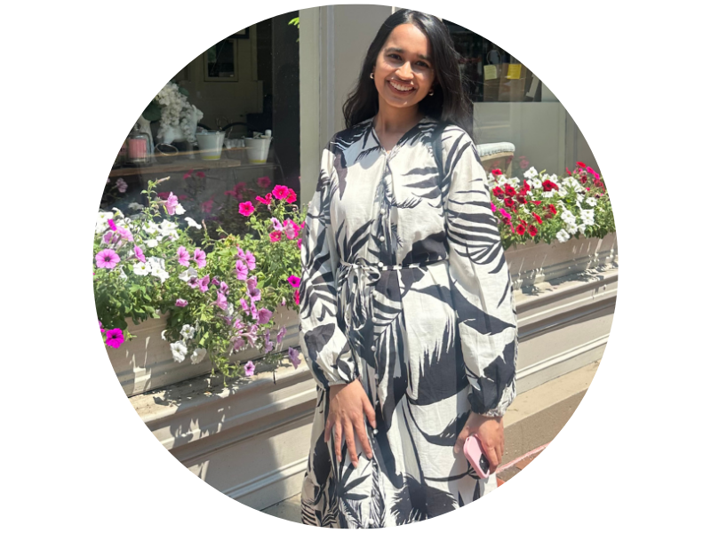 Photograph: SHAEERA RAHMAN, a young woman, in a patterned dress stands by colorful flower boxes outdoors.
