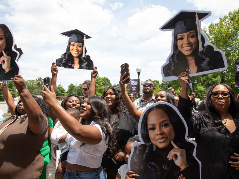 Crowd holds large cutout photos of smiling female graduates in caps and gowns outdoors.