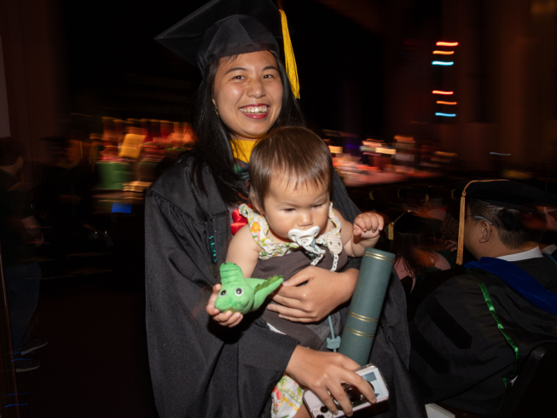 Smiling graduate in cap and gown holds a baby with pacifier, green toy, and a diploma.