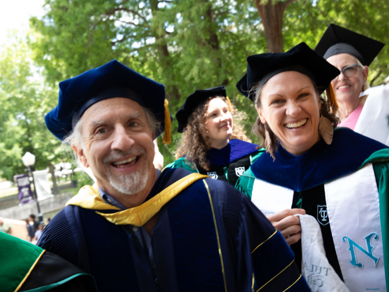 Photograph of happy graduates in blue and green academic regalia smiling outdoors.