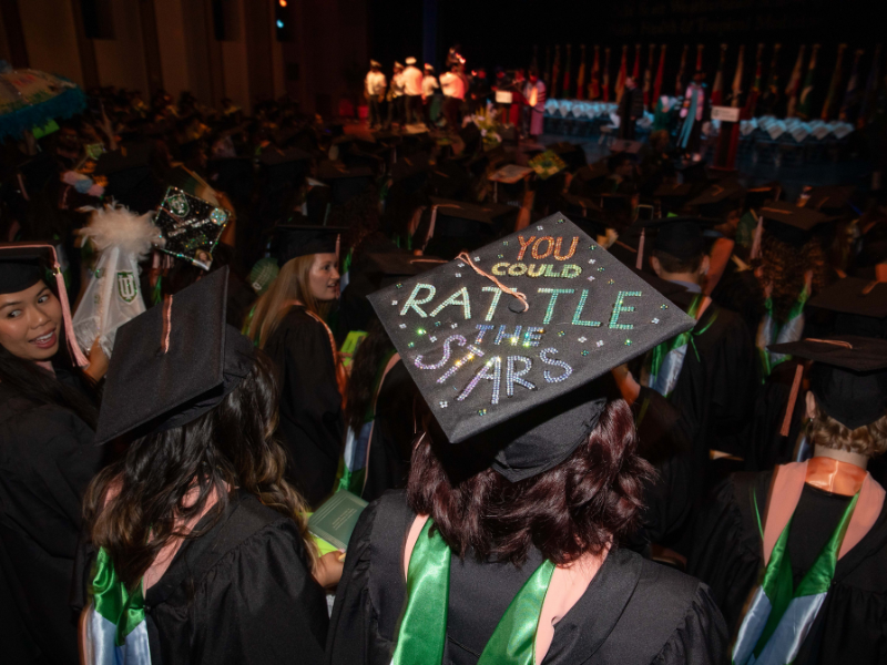 A graduate's cap is brightly decorated with a colorful inspirational quote, surrounded by other graduates.