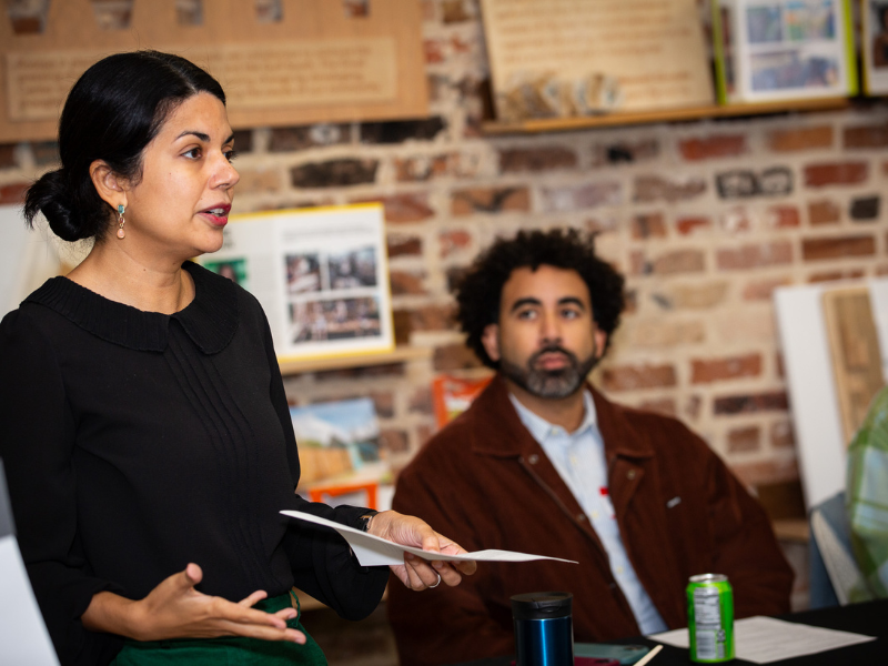 Melissa Fuster speaks, gesturing; man in brown jacket listens in background.