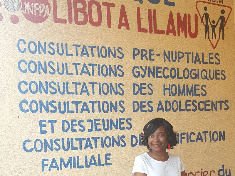 Alumna Gloria Kalong, MPH, standing in front of a hand painted sign at a health clinic in DRC. The sign lists the services provided at the clinic. 