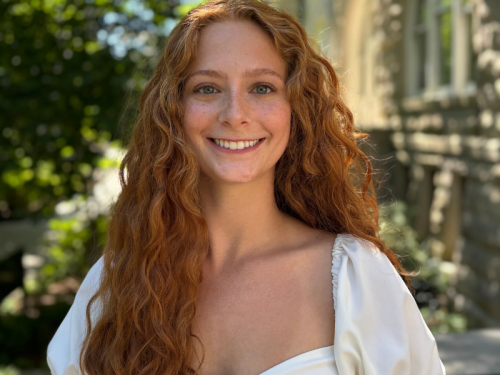 Ava Christiansen , with long red curly hair, wearing a white blouse and smiling at camera, standing outside Gibson Hall on Tulane University's uptown campus