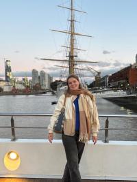 Maddy Williams smiling by a river at sunset, tall ship and city buildings in the background.
