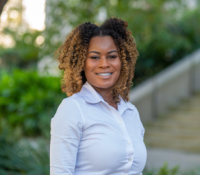 Alicia Williams, smiling, with curly hair in a white shirt outdoors.