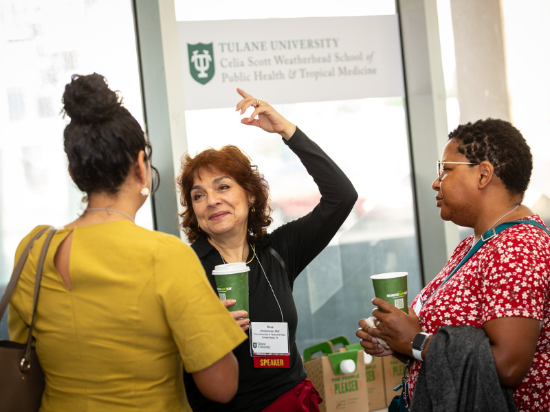 Three women chat, one gesturing, with the Tulane University logo in the background.