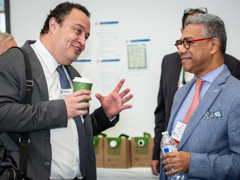 Two smiling men, one gesturing with a coffee cup, the other listening with a water bottle.