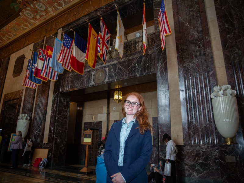 Red-haired student in glasses and suit smiles in a grand hall with international flags.