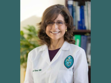 Smiling woman with glasses and brown hair wearing a white lab coat with an emblem.