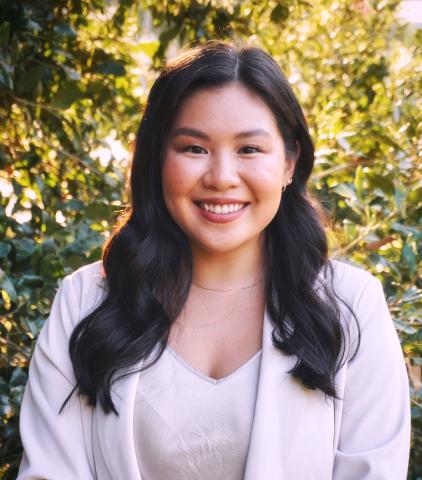 Smiling young Asian woman with dark hair in a blazer, green foliage background.