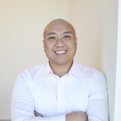 Smiling bald Asian man in a white shirt, arms crossed, leaning against a wall.